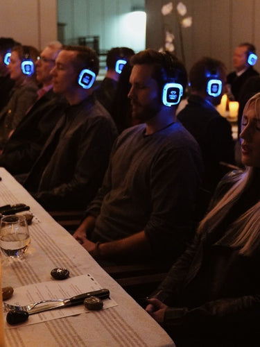 Indoor evening event featuring participants wearing advanced sound healing headsets, seated at elegantly arranged tables with no food, eyes closed in deep focus, experiencing a custom immersive sound healing session designed to elevate creativity, focus, and wellbeing for teams and leaders