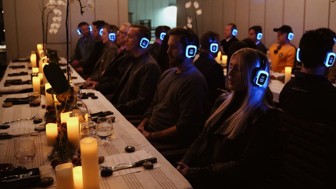 Indoor evening event featuring participants wearing advanced sound healing headsets, seated at elegantly arranged tables with no food, eyes closed in deep focus, experiencing a custom immersive sound healing session designed to elevate creativity, focus, and wellbeing for teams and leaders